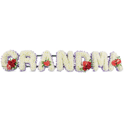 This image shows a beautifully composed floral lettering tribute spelling out GRANDMA in large, capital letters, the sort of bespoke funeral display often seen at services around Highgate and the nearby crematorium. Each individual letter is meticulously built from dense clusters of pure white cushion chrysanthemums, arranged so closely that they form a smooth, plush surface with a gentle, rounded texture. The white is softened further by a narrow trim of pale purple ribbon and backing that outlines each shape, giving the arrangement a subtle frame and a feeling of quiet serenity. Along the dark green metal framework that links the letters, the florist has introduced striking red floral highlights that punctuate the design. On the leading G, a lush group of deep red roses sits beside a single variegated red-and-white calla lily, its elegant, fluted bloom adding sophistication and movement. The first A carries a glossy, heart-shaped red anthurium lily at its base, like a small, shining emblem of devotion. At the centre of the name, the M is adorned with another cluster of red roses and a second red anthurium, drawing the eye towards the middle of the tribute. Finally, the last A holds one more red anthurium, echoing the earlier accents and providing a gentle visual rhythm from left to right. The overall mood is reverent yet tender, combining the innocence of white flowers with the rich symbolism of red. It is a thoughtful choice for families in Highgate, Archway, or Hampstead seeking a clear, loving way to honour their grandmother's memory.