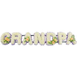 Against a crisp white background, a freestanding floral arrangement spells the word GRANDPA in bold, uppercase letters, each one carefully created from fresh, white flowers. The surface of every letter is densely covered with compact white chrysanthemums or similar small blossoms, their petals overlapping in rich layers that give a soft, almost velvety appearance. These flowers sit so closely together that the letters look solid and continuous, with no gaps, conveying both strength and delicacy. Around the outer edge of each character runs a narrow border of soft lavender or pale purple blooms, providing a gentle outline that enhances the readability of the word and introduces a tranquil, cool-toned accent. Woven into the white and lilac base are small yet significant clusters of peach roses and greenery. On the G and D, groups of two or three roses are nestled among slender white lily-like buds and deep green foliage, creating focal points of warmer colour. The first and final A each display a single peach rose with tidy sprays of green leaves, offering balance and a sense of quiet symmetry from one end of the tribute to the other. All seven letters are aligned along a thin, dark support bar, indicating a sturdy framework, likely metal, designed to stand during a funeral or memorial gathering. Viewed as a whole, the arrangement suggests the work of a dedicated florist in Highgate, perhaps prepared for a service close to Highgate Village or near the pathways of Hampstead Heath. It feels like a calm, dignified expression of remembrance for a grandfather, blending classic white funeral flowers with thoughtful accents that speak of family affection and cherished memories.