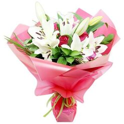 A romantic bouquet of white lilies and red roses is photographed against a simple white backdrop, the design evoking the refined yet slightly bohemian charm of Highgate's leafy streets. At the heart of the composition, several white Oriental lilies open into generous trumpets, their petals smooth, softly curved, and almost luminous, framing deep centres with dark, cocoa-brown anthers and slender pistils. Some lilies are caught mid-bloom, their petals not yet fully unfurled, while a few remain as tall, smooth buds with pale green casings, hinting at ongoing blossoms that will continue to open once the bouquet reaches a home near Highgate Village. Between the lilies, clusters of deep red rosebuds nestle closely, their petals tightly spiralled and velvety, adding richness, depth, and an unmistakable note of romance. The roses sit against lush, dark green foliage that creates a full backdrop, mixing broad, lustrous leaves with thin, arching blades that bring vertical lines and gentle movement, almost like the gentle sway of trees edging Hampstead Heath on a breezy day. Surrounding the flowers, a generous wrapping of bright pink, glossy cellophane is gathered and styled into soft, rippling folds that fan outward, catching light and reflections to create a festive, celebratory aura. At the base, a narrow lime green ribbon is tied with precision, its satin finish and fresh colour offering a playful counterpoint to the deeper tones above. The bouquet feels both timeless and modern, making it a fitting choice for anniversaries, heartfelt thank-you gifts, or same-day flower delivery throughout Highgate to mark life's joyful, intimate moments.