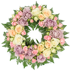 Captured against a plain white backdrop, this round funeral wreath presents itself as a serene, floral circle, thoughtfully composed of pastel roses and deep green fern foliage. The design feels intentionally full and opulent, a completely covered ring of blossoms where no structural base shows through. Velvety ivory and cream roses dominate the wreath, their fully opened petals unfolding in soft spirals, creating gentle shadows in their centres. Interspersed among these are sprays of blush pink roses, some still tightly furled, others beginning to open, adding a sense of time and continuity-buds, half-open flowers, and mature blooms living side by side. Warming the palette are soft peach and buttery yellow roses, while the inclusion of dusty lavender and muted mauve tones introduces a beautifully restrained, almost vintage elegance, well suited to a farewell in Highgate or nearby Crouch End. Behind and between the flowers, rich dark green fern fronds curve around the circle, their slender, feathered leaves creating a textured, woodland-like base reminiscent of the shaded paths of Queen's Wood. The even, diffused lighting allows the gentle colour transitions to be seen clearly, from cool cream to warm peach and into romantic mauve. The perfectly round form symbolises eternity and the cycle of life and remembrance, making the wreath an appropriate tribute for a funeral chapel, graveside, or crematorium service. As a whole, the arrangement feels composed yet deeply human, a carefully crafted expression of sympathy and enduring love that might be chosen by families across Highgate who wish to commemorate someone dear with quiet beauty and heartfelt respect.