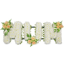 The image shows a refined sympathy floral arrangement spelling out MOM in bold, upright letters, composed with the care you might expect from a long-standing Highgate florist. Set against a clean, bright background, the word is instantly readable thanks to the dense covering of white spray chrysanthemums that form each letter. Every chrysanthemum head is small and neatly rounded, their petals overlapping into a perfectly even layer, creating a plush, almost quilted effect. The cool white of the flowers reflects the light softly, giving the tribute a calm, contemplative presence, as though it is already resting quietly in a chapel by Highgate Hill or prepared for a farewell at a North London crematorium. Just visible along the edges is a slim frame of dark green-likely foliage or ribbon-that traces the outlines of the M, O and M, refining their shape and adding subtle contrast. Across this gentle white surface are three thoughtfully arranged clusters of peach and apricot spray roses. One cluster sits at the top left of the first M, another is tucked low into the base of the central O, and the third rests on the upper right of the final M, creating a rhythmic balance of colour. Each cluster mixes open rose blooms with buds at various stages, surrounded by delicate fern, small sprigs of filler foliage, and slightly broader leaves, adding movement and a hint of garden freshness. The restrained palette and precise craftsmanship make this a fitting tribute for a mother, conveying love, respect and gratitude without overwhelming the quiet dignity of the occasion, whether displayed by the coffin or placed near the front during a celebration of life in Highgate.