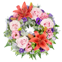 This photograph presents a circular sympathy wreath laid flat on a pure white surface, making the colours and textures of each bloom stand out with subtle elegance. The wreath is generously filled, forming a complete, unbroken ring that instantly suggests continuity and the ongoing nature of love and memory. Two large coral-orange Asiatic lilies command attention, one near the top and the other towards the lower section of the circle, their petals wide-spreading and gently curled at the tips, with prominent stamens adding a touch of drama. Surrounding these focal lilies, clusters of roses in shades of blush and lavender-pink fill the form, their velvety petals spiralling out in soft layers that convey warmth and affection. Interwoven with the roses are lisianthus blossoms, some a delicate pale pink and others a rich purple, their ruffled, cup-like shapes introducing a sense of movement and texture. Cool blue-grey eryngium thistles appear intermittently, their spiky forms and subtle metallic sheen adding architectural interest and a modern note. Wisps of tiny lavender-blue filler flowers, likely statice, drift through the spaces between larger blooms, creating an airy, mist-like effect. The base of the wreath is a robust bed of mixed greenery, with silvery eucalyptus, feathery ferns, and other textured leaves overlapping to form a deep green frame. A few closed lily buds rest amidst the foliage, hinting at blooms yet to open. Carefully folded bright green leaves near one lily suggest the practiced hand of a professional florist, such as one at Flowers Highgate preparing tributes for services along Highgate Hill or around the quiet streets near Highgate Wood. The overall impression is of calm beauty, respect, and heartfelt condolence.