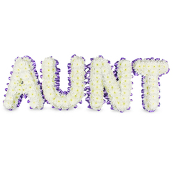 As a florist attentive to the responsibilities of remembrance, I describe this memorial piece as a clear, personal message: the letters AUNT rendered in bold, uppercase form and composed entirely of pristine white spray chrysanthemums. Each small bloom contributes to a textured, almost velvety surface, petals layered so densely that the eye reads a continuous plane of white interrupted only by the soft, pale-yellow centres peeking through like steady, gentle lights. A deep purple satin ribbon frames every letter with careful pleating and a slight ruffle, the fabric's sheen catching soft overhead light and providing a graceful border that lifts the whiteness away from the background. Beneath the letters, faint grey shadows indicate the three-dimensional frames and the way the setup sits away from the surface, giving the tribute quiet weight and presence. There is a faint, fresh scent of green stems and morning dew - subtle, dignified - and the arrangement's proportions feel steady and consoling, as if offering support in physical form. Created with the sensibility we bring to families in and around Highgate Village and along North Hill, this composition reads as both a public statement of loss and a private symbol of love. It is the sort of bespoke tribute many select for a funeral or a memorial at nearby chapels, where the white chrysanthemums and purple trim provide a tone of respect, warmth, and enduring remembrance without excess.