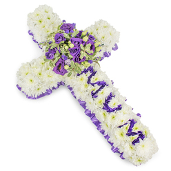 This cross-shaped sympathy tribute is photographed against a clean white backdrop so the craftsmanship is immediately legible: the cross itself is entirely covered in tightly placed white chrysanthemum blooms, creating a soft, pillowy texture that suggests serenity and repose. Encircling the edge is a ruffled purple satin ribbon with a subtle sheen that defines the silhouette and lends a formal, graceful border. Down the vertical shaft the personal name MUM is formed from clusters of small purple blooms-likely eustoma or compact spray chrysanthemums-carefully set to read clearly from a short distance, a private message rendered in floral form. The central focal spray at the crossing is generous and layered: deep violet roses with velvety petals combine with lighter lavender lisianthus or eustoma and sprigs of variegated greenery, whose pale-edged leaves and darker centres bring depth and a touch of gentle contrast. Tiny green berries and delicate foliage echoes through the arrangement, adding texture and quiet movement. The cool palette-crisp white paired with varying purples-evokes dignity, peace, and a restrained sorrow, and there is a soft, fresh fragrance reminiscent of late-spring gardens after rain. As an experienced florist working in Highgate, I picture this piece being chosen for a chapel service near Highgate Village or a quiet graveside at the local cemetery; it is composed to read well in both close-up photographs and house-of-worship settings, offering a tangible expression of enduring affection while remaining calm, composed, and deeply respectful.