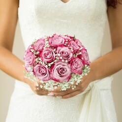 This evocative image embodies the beauty and artistry found in Highgate's wedding traditions. A hand-tied bouquet sits at the heart of the photo, exuding an understated elegance shaped by expert local hands. The roses rise in a seamless blend of pale pink and deep magenta, their petals arranged in a perfectly rounded sphere that glows with romantic warmth. Wisps of baby's breath, white and cloud-like, thread between the blooms, capturing the dewy brightness of a Highgate spring morning. The bride's hands-skin warmed by time outdoors on Hampstead Lane or Highgate West Hill-cup the bouquet with a sense of calm assurance. Her nails, tastefully manicured in a light shade, rest against her dress's distinctive lace overlay and delicate V-neckline, a reminder of the classic style that resonates at Highgate's most cherished venues. The bouquet seems to float, radiating both serenity and excitement, as if imbued with all the hope and promise of a new beginning. Designed by Flowers Highgate, this arrangement is meant for couples who desire a bouquet as memorable as their love story, crafted with devotion and delivered with pride to every corner of the local community.