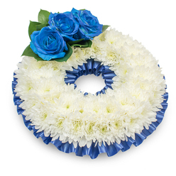 Photographed from a modestly raised vantage point against a clean white backdrop, this wreath presents a composed circle of remembrance. The dominant band is crafted from tightly packed, snow-white chrysanthemums whose innumerable feathery petals create a plush, cloud-like texture and a gently domed silhouette that throws a soft shadow on the surface below. Nestled at the upper left, three luminous blue roses provide a striking focal accent; their petals are densely furled and velvety, the deep sapphire tone intentionally bold against the chrysanthemum field. A handful of glossy dark-green leaves surrounds the roses, offering contrast in both colour and texture and giving the cluster a natural, leafy cradle. Both the inner and outer perimeters of the wreath are edged with a pleated cobalt satin ribbon that mirrors the roses' blue - its ruffles and sheen lend discipline and polish to the design while emphasizing the wreath's circular geometry.Beyond visual detail, the arrangement conveys quiet sensory hints: the soft, powdery scent of the chrysanthemums, the tactile softness of petals, and the cool glide of satin ribbon when touched. As a florist in Highgate, I imagine this piece placed with dignity at a memorial service near Highgate Village or laid at a graveside in Highgate Cemetery; its palette and form read as traditional yet personal, intended to console and honour. The overall effect is restrained, noble, and thoughtfully composed - a tribute that balances classic symbolism with a contemporary, local florist's attention to finish and proportion.