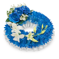 Presented against a clean white backdrop, this memorial wreath is a statement of serene craftsmanship from a Highgate florist's hands: a dense, circular base of vivid royal-blue chrysanthemums forms a rich, textured ring whose tiny, spidery petals create a tactile, cloud-like surface. The inner circumference is softly lined with pleated white satin ribbon; its glossy folds catch the light and offer a gentle punctuation between the deep blue flowers and the empty central space. At the upper-left quadrant a cluster of three full, velvety blue roses commands attention, their petals unfurling in layered sapphire tones and surrounded by delicate clusters of white gypsophila and small, dark green foliage that provide subtle contrast and depth. Flowing from the mid-left and arcing toward the lower-right is a cascade of ivory-white orchids - probably Phalaenopsis or Dendrobium - their broad, silky petals and sculpted lips creating an ethereal sweep across the wreath. Interspersed among those blooms are fresh green orchid buds, some closed, some poised to open, suggesting continuity and quiet hope. The composition reads as dignified and contemplative, the textures ranging from velvety to crisp satin to the airy filigree of baby's breath. Flowers Highgate prepares and delivers similar tributes locally; for families in Highgate Village, Archway and surrounding streets we can arrange careful delivery and siting to honour a loved one with understated beauty and respect.