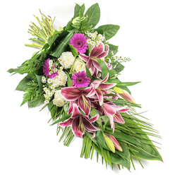 A refined funeral sheaf, photographed on a bright white background, stretches diagonally like a gentle arc of colour and foliage. From the neatly bound stems at the top left, the eye is drawn down through layers of blossoms to a tapered finish of cascading leaves and soft fern fronds at the bottom right. At its heart are striking Stargazer lilies, their velvety petals saturated with deep fuchsia-pink and magenta shades, dotted with burgundy speckles and edged in pure white. Some flowers open fully to reveal their stamens, while others remain in bud, offering a quiet suggestion of unfolding remembrance. Surrounding the lilies are clusters of creamy white roses and plush, ruffled white carnations, which add volume and an almost cloud-like softness. Bright magenta gerbera daisies are woven throughout the design, providing strong, circular accents and a sense of warm affection. Fine white filler flowers, akin to tiny daisies, intermingle among the larger blooms, creating a lace-like veil of detail. The greenery is lush and considered: broad, glossy dark leaves ground the arrangement, feathery ferns add natural lightness, and long, blade-like leaves sweep outward to enhance its elongated form. The overall mood is both solemn and reassuring, making this spray an apt choice for funeral or memorial services around Highgate, whether for a ceremony near Highgate Wood, a chapel close to Archway Road, or a quiet family farewell at home. It feels like the work of a dedicated local florist, carefully balancing colour, texture, and symbolism to convey sympathy, respect, and enduring love.