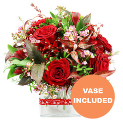 A rich, romantic bouquet is pictured against a clean white backdrop, its colours and textures carefully balanced to form a full, rounded arrangement. Deep red roses are the undeniable stars here, their velvety petals unfolding in spirals that range from tightly furled buds to more open, inviting blooms that seem to breathe warmth and emotion. Nestled around them are smaller deep crimson flowers, akin to alstroemeria or similar multi-petalled varieties, whose slim petals and speckled faces give the design a natural, garden-inspired detail. Threaded through this sea of red, fine sprigs of tiny pale pink and white blossoms add a gentle, airy contrast, each miniature flower catching the light and bringing a sense of softness, like a whisper between more intense notes. The whole bouquet is framed with a generous collar of greenery: glossy dark leaves, lighter green tips, and more delicate, feathery textures combine to make the arrangement feel lush and well-rounded. Among the foliage, burgundy and reddish-brown leaves with subtle patterning introduce a deeper, more dramatic tone, harmonising with the roses and adding an elegant edge that would suit a stylish Highgate residence near Hampstead Lane or North Road. The stems are gathered neatly in a clear rectangular glass vase with smooth edges, allowing the viewer to see the clean water and meticulous conditioning beneath. Around the vase, a red ribbon decorated with a repeating pattern of white heart outlines offers a gentle nod to romance without overwhelming the design. In the lower right corner, a bright orange circular overlay carries bold white uppercase text reading "VASE INCLUDED," making it clear that this Highgate bouquet is delivered ready to display, ideal for anniversaries, intimate dinners, or thoughtful surprises.