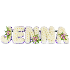 This personalised memorial spells the name JENNA in five substantial, three-dimensional letters, each built from a dense blanket of small, creamy white chrysanthemums or similar florets. The result is a soft, cloudlike texture that looks almost cushiony to the touch, while a slender ribbon or a line of tiny purple blooms frames the interior edges of every letter, providing refined definition and a thoughtful colour note. We've added three modest accent posies - blush-pink and lavender spray roses with a few cream or pale yellow roselets and wiry green fern fronds - positioned on the curved arm of the J, the centre of the first N, and the upper sweep of the A, so the arrangement reads as both sculptural and intimately floral. Dark, unobtrusive supports run horizontally behind the letters, indicating the arrangement is built to be freestanding for placement beside a casket or against a memorial screen. The subtle scent of the roses blends with the clean, green aroma of the ferns, while the chrysanthemums lend a restrained, comforting presence. As a Highgate florist with experience placing tributes at local services - from the chapel near Highgate Village to quiet moments at Hampstead Heath's edges - I picture this namepiece offered to families who want a personal, elegant farewell. Crafted with attention to proportion and readability, it honours a name with calm dignity and considered floral detail.