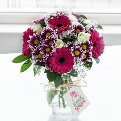 A lush, professionally arranged bouquet is showcased against a softly diffused white backdrop, as though placed on a bright Highgate windowsill on a calm morning. At the heart of the arrangement, bold gerbera daisies in intense fuchsia and magenta seem to radiate energy, each flower head perfectly formed with a deep, almost black centre that adds drama. Around them, bicolour chrysanthemums in rich purple and white contribute a lively, almost playful rhythm, their many petals edged in strong colour that gradually lightens toward bright yellow centres. Between these statement blooms, clusters of creamy white roses are tucked in with care; some are fully open, revealing layered petals that look almost satin-smooth, while others remain in bud, hinting at beauty still to come. Fine sprays of white baby's breath weave throughout the bouquet like a soft cloud, giving the composition gentle movement and a romantic, airy quality. Dark, glossy green leaves and foliage support the flowers, creating a lush base and adding contrast that makes the pinks and purples sing. The stems are gathered into a clear, jar-style glass vase whose textured surface catches the light, and a rustic twine bow is neatly tied around the neck. A small white tag with a simple pink blossom illustration dangles from the twine, suggesting a personal note from someone sending love, congratulations, or a get-well wish. The overall mood is joyful and uplifting, perfect for brightening a flat off Highgate High Street or welcoming guests in a home near Hampstead Heath, and it quietly reflects the skill of a local florist who understands how to balance colour, texture, and sentiment in a single arrangement.