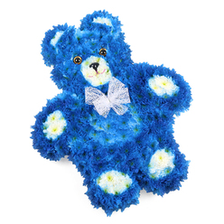 A floral teddy bear lies diagonally across a white background, its sculpted body formed entirely from densely packed blossoms in deep blue and soft cream. The main structure of the bear is created with royal blue flowers, likely small chrysanthemums or similar button blooms, placed so closely together that they resemble plush fur, every curve and contour of the bear's body clearly defined. Cream-coloured flowers, with a subtle buttery warmth, highlight the muzzle, the inner circles of the ears, and four rounded paw pads, breaking up the intense blue and adding gentle definition to its familiar teddy shape. The face is particularly expressive: two round, glossy eyes in dark brown or amber catch the light, while a neat black triangle nose and a fine, curved mouthline suggest a quiet, comforting smile that feels protective and calm. A delicate white bow in a mesh or organza fabric is tied neatly around the bear's neck, its sheer, slightly ruffled texture introducing a touch of elegance that lifts the design beyond simple novelty. The overall composition is clean and balanced, with the bright white backdrop ensuring that the colours and textures of the flowers are clearly visible. From the perspective of a Highgate florist, this kind of floral sculpture is often chosen for tender occasions involving children or babies-perhaps a memorial held in a church near Highgate Village or a farewell gathering close to Parliament Hill-where families seek a tribute that speaks of innocence, love, and cherished memories. The piece radiates both sorrow and solace, like a cuddly toy transformed into a living, fragrant symbol of remembrance.