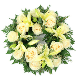Viewed flat from above on a plain white surface, this elegant funeral wreath brings together a soft palette of cream and pale yellow that speaks of calm and consolation. At its heart sit about fifteen to twenty ivory roses, their petals tightly spiralled and catching a gentle, diffuse light to appear almost luminous. Interspersed among them are generous, trumpet-shaped lilies in a tender butter-yellow; some petals are unfurled in graceful arcs, revealing delicate stamens, while others remain in poised bud form - a deliberate mix that introduces movement and a quiet hope of renewal. The blooms are anchored by a generous base of deep green foliage: delicate fern fronds lend a feathery, almost cloud-like edge, while broader, rounded leaves resembling eucalyptus add shadowed tones and structural depth. The overall composition is densely packed yet airy, balanced and symmetrical in its circular form so that each flower reads clearly from above. There is a soft, layered scent - powdery rose undercut with the green, faintly citrus notes of lily - that suggests tenderness more than ostentation. Designed as a memorial or sympathy tribute, this wreath conveys purity, respect, and enduring remembrance. Prepared locally with care for families across Highgate - from the lanes around Highgate Cemetery to the paths of Hampstead Heath - and available through Flowers Highgate for discreet local delivery, it is a considered choice for a funeral service or a graveside homage.