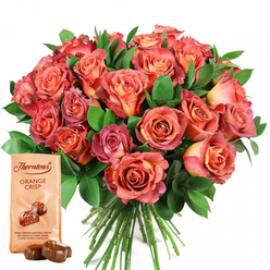 Every facet of this thoughtfully styled floral ensemble, arranged by a seasoned florist with an appreciation for Highgate's timeless charm, speaks to the interplay of colour, texture, and heartfelt intent. The bouquet is a generous gathering of roses in full bloom, each petal showcasing gradations that move from gentle cream and buttery peach at their centres to luminous coral and velvety pink at the edges. Soft daylight glances off the petals, highlighting their velvet finish and subtle ombre, creating a sense of movement akin to a gentle breeze through Highgate Wood. Encircling the heart of the arrangement is a lush frame of dark fern and eucalyptus, their cool greens a deliberate contrast that makes the rose colours glow. To the left in the foreground, a carefully placed pouch of Thorntons Orange Crisp chocolates lends a note of sweet indulgence, its pale wrapper featuring the familiar logo above an illustration of glossy chocolate truffles-a visual promise of zesty orange layered within milk chocolate. Two actual truffles rest before the package, their sheen echoing the inviting luxury within. This union of fresh blooms and fine chocolate evokes celebration and affection-perfect for birthdays or anniversaries in Highgate. It's a tribute to the artistry and tradition found in every bouquet crafted for the N6 community.