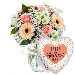 This Mother's Day scene feels like a quiet celebration captured in still life, styled against a clean white background that could easily be a Highgate kitchen wall in soft morning light. A clear glass vase, uncomplicated and cylindrical, stands firmly at the centre, its neck circled with a light brown twine bow that introduces a touch of rustic charm. From the vase rises a lush bouquet of spring flowers in gentle pastel shades. The roses, in peach-pink tones, are fully open and layered, each petal curling outward in a neat spiral that conveys elegance and tenderness. Nestled among them, large peach gerbera daisies, with velvety petals radiating from dark brown centres, bring a cheerful, modern sparkle. Smaller white chrysanthemums, rounded and compact, peep out with subtle pale green centres, adding texture and visual rhythm. Airy sprays of white gypsophila wind their way through the larger blooms, creating a misty, romantic backdrop that gives the arrangement a graceful, floating quality echoing walks along Highgate's tree-lined streets. Emerald green foliage provides contrast and structure, cradling the softer hues. Tucked behind the flowers, a small white card with blurred pastel writing suggests a personal Mother's Day message, private yet central to the gift. In front of the vase to the right, a heart-shaped foil balloon nestles close. Its white surface is sprinkled with glimmering gold and silver dots, like confetti frozen in mid-air. At its centre, a peach heart contains the words "HAPPY Mother's DAY" in black, with the word "Mother's" written in elegant calligraphy that draws the eye. The overall composition feels warm and heartfelt, a ready-to-deliver bouquet and balloon pairing that would suit a Highgate home filled with family stories and gentle appreciation.