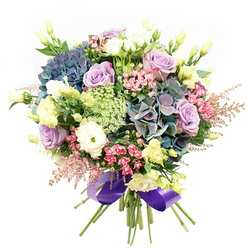 This lavish bouquet, meticulously crafted for Highgate's discerning community, stands out against a luminous white backdrop. Sweeping hydrangea heads in dusky indigo, velvety teal, and soft violet evoke the evening shades drifting across the Hampstead Heath skyline. Soft lavender roses and creamy white blooms open at the centre, their gently curled petals a testament to timeless romance and celebration, perfect for birthdays or anniversaries marked in Highgate's historic homes. White ranunculus is tightly packed for contrast, underscoring the arrangement's plush texture. Tall, slender eustoma (lisianthus) in buttery yellow hues rise gracefully, mingling with vivid fuchsia-pink fillers and airy sprays of blush astilbe that trail like flowering vines down Bisham Gardens. Interwoven sprays of light green and white, reminiscent of Queen Anne's Lace, add depth and brighten the bouquet with intricate, almost luminous detail. Glossy, deep green leaves set the blooms aglow, while the neatly gathered stems are tied in a lustrous royal purple satin ribbon-a flourish that elevates the bouquet's artistry and nods to Highgate's creative heart. Perfectly balanced between classic style and modern vibrancy, this arrangement delivers joyful sophistication to homes, events, or thoughtful gifts throughout N6.