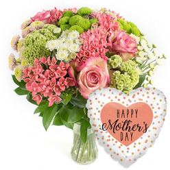 This photograph captures a joyful Mother's Day bouquet and balloon combination, styled with the understated elegance you often see in Highgate homes. On the left, a clear glass vase holds a lush, dome-shaped arrangement, its stems neatly aligned in the water. The bouquet is led by several large pink roses, each bloom opening into velvety spirals, with a slight deepening of colour along the petal edges that adds depth and romance. Threaded closely around them are clusters of coral-pink blossoms, much like kalanchoe or compact spiky florets, creating pockets of warm, sunlit colour. Lime-green button chrysanthemums pop between these clusters, their rounded, almost cushion-like heads and other chartreuse textured blooms introducing a fresh, contemporary twist that feels in tune with spring walks through Waterlow Park. Tiny white spray chrysanthemums, evocative of daisies, are dotted through the design, their crisp petals and yellowish centres bringing brightness and balance. Lush, dark green foliage cradles the entire arrangement, adding structure and a sense of abundance. To the lower right of the vase, a festive heart-shaped foil balloon leans into the composition. The balloon's surface is predominantly white or pale silver, decorated with scattered metallic spots in gold, silver and peach, giving the impression of floating confetti. At its centre sits a solid peach heart, inside which the words "HAPPY Mother's DAY" are printed in stylish black lettering, combining bold capitals with elegant script. The overall feeling is one of gratitude and celebration, the kind of floral gift you might send to a mother in Highgate Village, along Archway Road, or near Highgate Cemetery, turning an ordinary living room into a small moment of festivity and love.