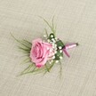 A single pink rose boutonniere rests on a softly textured, pale beige linen cloth, captured from a gently elevated angle so that every detail of the florist's work is visible. The rose itself is semi-open, its velvety petals spiralling outwards from a tight pastel pink heart into slightly deeper, more romantic blush tones at the curled outer edges, suggesting a bloom cut at exactly the right moment. Clusters of creamy white baby's breath surround the base of the rose like a delicate cloud, each tiny blossom adding airiness and a sense of classic bridal elegance. Fine, grass-like strands of light, fresh green foliage fan out behind the flower, giving the piece a feeling of movement, as if a soft breeze from Hampstead Heath had just brushed through it. A broader, darker green leaf is folded neatly around the stems, creating a smooth cradle that keeps the composition tidy and structured, much like a well-pressed lapel on a suit worn to a Highgate Village wedding. The stems are carefully wrapped in a satin ribbon that shifts from lavender-pink to deeper magenta-purple, the sheen of the fabric catching the light and adding a gentle hint of luxury without overpowering the softness of the rose. The understated linen background, reminiscent of a natural table runner you might see in a Highgate townhouse or café off Highgate High Street, allows the colours to stand out while also hinting at a relaxed, modern London style. The overall effect is tender and romantic, a small but meaningful floral detail ready to be pinned to a groom, groomsman, or prom guest for a day of celebration.