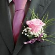 There's a gentle lyricism woven into this Highgate buttonhole-a design that speaks to romance and anticipation in every exquisitely arranged element. At its core lies a rose in cool blush pink, petals soft and unfurling, evoking notes of springtime air drifting through the local church gardens. Encircling the bloom, pearly clusters of baby's breath contribute an almost magical lightness, lending a dreamy softness that highlights the rose's elegant silhouette. Textural, slender green leaves and broader, dark green accents curve quietly behind, suggesting both woodland beauty and urban refinement. Secured with a delicate lilac ribbon, the boutonniere's artistry is mirrored in the vivid pink tie and gently patterned grey shirt beside it. This ensemble, finished by the tailored charcoal suit lapel and a precise buttonhole, is more than a floral accent-it's a symbol of cherished moments and community celebration in Highgate, radiating understated grace and a sense of hope on life's special occasions.