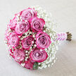 A hand-crafted bouquet of roses and baby's breath rests on a pale, textured cloth that resembles soft linen, the neutral backdrop allowing every pink and white detail to stand out, much like a bridal bouquet laid out in a quiet Highgate townhouse before the ceremony. At the core, about a dozen roses create a lush, rounded cluster. Their petals transition gracefully from muted, dusty lavender-pink near the centre to intense fuchsia and deep, almost berry-toned magenta along the outer rims. Each flower is full of layered, velvety petals, some still tightly closed in a compact spiral, others beginning to loosen and curl outward, giving the design a sense of unfolding romance. Misty white gypsophila, or baby's breath, fills the spaces around the roses, spilling out to form a soft, cloud-like border. The tiny clustered blossoms lend the bouquet an airy, delicate appearance, tempering the boldness of the rose colour and adding an unmistakably bridal charm. Here and there, hints of dark green foliage peek out beneath the blooms, grounding the palette and evoking fresh garden growth like you might see in nearby Highgate Wood. The stems are neatly aligned and bound together at the bottom right, wrapped in a smooth lilac satin ribbon that gleams gently under the light. Wound around this handle is a fine spiral of small white pearls, referencing traditional wedding accessories and adding a note of refinement. The soft, even lighting and the off-white fabric background emphasise the bouquet's colour gradient and texture, making this arrangement perfect for weddings, anniversaries, or same-day flower delivery across Highgate for those wanting to send a message of graceful, enduring love.