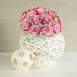 In this image, a modern wedding arrangement exudes quiet luxury and considered design, much like the atmosphere of a refined Highgate home. At the centre sits a perfectly shaped mound of pink roses, carefully massed so that the blossoms form a smooth, dome-like silhouette. The petals are densely layered and velvety, blending from soft, powdery pink near the heart of each flower into deeper, berry-toned edges that create gentle shadows and highlights across the dome. Tiny clusters of white baby's breath are tucked among the roses, forming a delicate, misty ring that outlines the base of the bouquet and adds feathery contrast to the lush petals. Cradling this composition is a distinctive white spherical container, crafted from interwoven strands that loop into a hollow orb. The open structure catches the light, sending slender lines of shadow onto the surface beneath and allowing glimpses of the stems and floral mechanics inside. Beside the vessel, two small angular ornaments rest with quiet confidence, their minimal shapes harmonising with the globe's contemporary aesthetic. The background is simple and luminous, so all attention remains on the interplay between the pink roses, white gypsophila, and the sculptural vase. The mood suggests a relaxed yet polished celebration-perhaps a civil ceremony followed by a gathering in a Highgate restaurant, or a chic reception in a home near Highgate Wood. As you imagine the scene, it's easy to sense the gentle fragrance of roses mingling with soft conversation, making this arrangement ideal for wedding tables, cake displays, or as a romantic focal point in photographs capturing love and commitment.