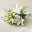 This fresh, ethereal arrangement rests on a pale beige linen cloth, lit by soft natural light that throws gentle shadows and reveals every subtle texture. Dominant, pristine white lilies stand tall with satin-smooth petals curling back to expose creamy centres and golden stamens flecked with tiny dark dots, while green, tightly furled buds promise further unfolding. Interspersed among the lilies, white lisianthus provide a delicate counterpoint: their petals edged with a faint lavender-purple blush, ruffled and layered so each blossom offers a different silhouette. Wisps of fine, fern-like greenery thread through the cluster, lending a weightless, meadow-like quality, and larger glossy leaves give the bouquet a rich green foundation. Small clusters of baby's breath are woven throughout, their minute white flowers acting like soft confetti that brightens the spaces between larger blooms. The stems are hand-tied and wrapped in a neat white satin ribbon at the right side, a tactile finish that speaks of careful florist craft. As a local florist, we picture this bouquet sitting on a café table in Highgate Village or brightening a mantelpiece after a stroll across Hampstead Heath; its scent is gentle, almost creamy, and the arrangement radiates calm celebration. Prepared with care by Flowers Highgate, it reads as an elegant option for intimate weddings, anniversary gifts, or a thoughtful gesture to bring quiet beauty into a Highgate home.