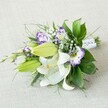 A hand-tied spray of white lilies and purple lisianthus lies diagonally across a soft, light beige fabric, the weave of the cloth subtly visible beneath the stems. The bouquet feels like it has just been set down on a table in a Highgate townhouse, still cool and dewy from conditioning, its petals catching gentle, diffused light. At the heart of the arrangement, several large white Oriental lilies are in full, elegant bloom, their wide, recurved petals touched with faint speckles and framing rich yellow stamens dusted in warm pollen. Nearby, plump chartreuse buds point outward, not yet opened, promising more fragrant flowers over the next few days. Threaded between the lilies, ruffled lisianthus blossoms in white with vivid purple edges create little pools of colour, the tones ranging from soft lavender to deeper violet that echo the dusky skies above Waterlow Park. Fine sprigs of delicate white gypsophila float throughout like tiny clouds, adding airiness and a romantic, bridal softness. Lush greenery wraps around the blooms, from broad, glossy dark green leaves that anchor the shape to finer, spiky fronds that add movement and a slightly wild, garden-picked feel, as though gathered from a Highgate Village garden after rain. At the narrow right end, the stems are neatly bound in a white satin ribbon, accented with subtle pearl-like beads that catch the light without overpowering the flowers, suggesting a bouquet intended for a wedding, an anniversary walk across Hampstead Heath, or a meaningful celebration at home. The overall impression is one of calm elegance, freshness, and quiet luxury, ideal for local Highgate flower delivery when you want something refined yet deeply expressive.