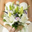 In the heart of Highgate, where cobblestone lanes meet sprawling gardens, this exquisite bridal bouquet captures the spirit of North London elegance. The photograph reveals a sumptuous composition, centered on luminous white Oriental lilies, their generous petals dappled with faint spots and cradling golden stamens. Nestled beside them are lisianthus, their soft, white blooms rimmed with gentle lavender and wisps of color, swirling among buds waiting to unfurl. Arching upward, royal purple veronica lends a sculptural contrast, while clouds of dainty baby's breath scatter a misty veil over the arrangement. The bouquet is anchored by a tapestry of foliage-broad, healthy leaves, unopened lily buds, and the tousled green of asparagus fern-mirroring the lush greenery found in Highgate's parks. Wrapped in the bride's hands with understated nail polish, the bouquet stands out against her softly blurred white strapless dress, radiating both intimacy and grandeur. Designed with local expertise, this bouquet is perfectly suited for Highgate weddings, echoing the romantic promise of each new day and ready for delivery to cherished celebrations along Pond Square or near Highgate Wood.