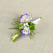 Pinned gently on neutral linen for the photograph, this boutonniere is a compact study in soft contrasts: two lisianthus blooms with crisp white faces and a subtle violet halo, set amid feathery greenery and fine, cloud-like sprays of baby's breath. The lisianthus petals appear almost silk-fine, curling at the edges with a tactile, velvety look, while clusters of grape hyacinth introduce a vivid blue-violet punctuation-tight, columnar spikes that lift the composition and add a lively spring energy. Pale, elongated buds thread through the arrangement, lending a sense of upward movement, and slim fern fronds fan out to create a woodland fringe that keeps the design feeling fresh and organic. The stems are tied with a tidy white satin ribbon whose sheen reads faint against the textured linen beneath; the overall scale is intimate, meant to sit lightly on a lapel or be tucked into a jacket pocket for a wedding, anniversary, or formal portrait. Photographed in soft, natural light, the colours retain a cool clarity-the whites clean, the greens deep and reassuring-evoking early-morning walks around Swain's Lane and the greener corners of Highgate Village. As a hands-on maker from Flowers Highgate, I picture this piece being chosen for its gentle scent, subtle visual dialogue with a suit, and the way small details can carry a day's memory.
