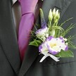 A close-up photograph captures the upper torso of someone dressed for a wedding or smart celebration in Highgate, with the focus drawn to a meticulously crafted boutonniere pinned to a deep charcoal grey suit jacket. The fabric of the jacket looks smooth and finely tailored, the lapel sharply defined, while beneath it a light grey checkered dress shirt introduces a soft pattern. At the centre of the image, a rich, lustrous magenta silk tie glows against the cooler greys, its knot perfectly aligned, echoing the sense of occasion one might feel heading to a ceremony near Waterlow Park or a reception just off Highgate High Street. Nestled against the lapel, the floral buttonhole is the true star: a cluster of white lisianthus flowers edged in vivid purple, their ruffled petals curling gently like painted brushstrokes around sunny yellow centres. Around them, several slender, unopened white buds hint at blooms still to come, while a slim spike of dark violet veronica rises gracefully, adding vertical interest and a slightly wild garden feel, as if freshly picked from a border in a leafy N6 back garden. Tiny white filler flowers reminiscent of baby's breath sprinkle lightness through the design, cushioned by mixed greenery-feathery fronds, smooth emerald leaves, and looping strands of decorative grass that bring a modern, sculpted touch. At the base, a small, immaculate white satin ribbon is tied into a neat bow, securing the stems and adding a traditional, bridal elegance. The lighting is soft and flattering, gently reflecting off the tie and the satin bow, creating an atmosphere of poised anticipation and quiet joy. It feels like the thoughtful finishing detail on a groom's outfit, lovingly arranged by an experienced Highgate florist for a day that will be long remembered.