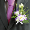 Nestled against the deep charcoal of a well-cut suit, this boutonnière has the calm assurance of something made by hands that know their neighbourhood-think Highgate Village flower markets and the cool air by Waterlow Park. Its focal bloom is a white lisianthus edged with a bright violet-purple, petals soft and slightly ruffled, a tactile centre that invites a gentle brush of the fingertip. Beside it a white dianthus with lively purple frills adds a playful, textured contrast; together they read like a small duet of refined colour. A slim spike of indigo veronica lifts the eye, while delicate grassy loops and glossy green foliage create a natural frame that moves with the wearer. Sprigs of gypsophila float like hints of light, adding air and a whisper of innocence; three pale lisianthus buds stand ready behind the main bloom, promising unfolding moments. A neat white ribbon secures the composition, clean and unassuming against the dark fabric, while the nearby magenta tie sets up a sophisticated, modern pairing. The boutonnière gives off a soft garden scent, fresh and reassuring over the course of a long celebration-weddings, anniversaries, or any milestone where detail matters. As prepared by Flowers Highgate, it reflects local craft: measured, elegant, and quietly distinctive-perfect for someone walking down Highgate Hill or pausing by the cemetery gates, carrying a small, fragrant emblem of care.
