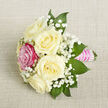 On a pale beige, subtly textured fabric that looks like fine linen laid out on a styling table, a petite bouquet is positioned diagonally, as if just placed down by a careful hand. Five ivory and creamy white roses form the main cluster, each bloom full and rounded, with petals unfolding in soft spirals that convey quiet luxury and freshness. Nestled among them, a single fuchsia-pink rose draws the eye, its saturated hue adding a confident pop of colour that plays beautifully against the otherwise gentle palette, reminiscent of the bursts of blossom that brighten Highgate's leafy residential streets in spring. Airy sprays of white baby's breath thread through the arrangement, their tiny blossoms creating a lace-like veil that lightens the overall feel and introduces a sense of delicacy and innocence. Around the flowers, deep green, tapered leaves give shape and balance, their smooth surfaces and defined edges providing contrast in both tone and texture. The stems are carefully gathered and wrapped in a bright pink satin ribbon, which forms a polished handle, smooth and slightly reflective under the soft, even lighting. Over this ribbon, rows of small, iridescent white pearls are arranged with precision, suggesting the piece is meant for a cherished occasion: perhaps a corsage for a Highgate School prom, a groom's clustered boutonnière, or a petite bridal bouquet. The whole composition feels intimate, romantic, and handcrafted, the kind of thoughtful floral detail one might choose for a meaningful celebration or an elegant dinner along Highgate High Street, where every element speaks of intention and care.