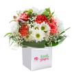 There's an infectious happiness about this bouquet, displayed in a sharp, modern white gift bag-like a burst of spring sunshine welcomed on Highgate's historic streets. Soft coral and vibrant red carnations, their petals exquisitely frilled, lean affectionately beside crisp, white daisies, each flower further brightened by vivid green-yellow centers. Billowing tufts of white baby's breath mingle playfully throughout, adding a soft, airy lightness that recalls the breezes drifting through Highgate Cemetery's ancient trees. Subtle hints of purple in the form of unopened buds appear at one side, delicately hinting at moments of gentle surprise and anticipation. The entire arrangement is circled by deep emerald foliage and glossy broad leaves, giving it a lush, rounded embrace that anchors every vibrant detail. The pristine white gift bag, with smooth ribbon handles atop, elevates the bouquet-making it perfect for celebratory birthdays, heartfelt anniversaries, or spontaneous joys shared with neighbours from Archway to Hampstead Lane. Suffused with freshness and crafted by skilled local florists, this floral ensemble encapsulates the delight of gifting and receiving flowers within the warm, storied community of Highgate.