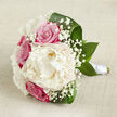 On a softly creased, light beige fabric that could be a linen runner on a Highgate townhouse table, a wedding bouquet is laid horizontally, inviting a lingering look. A single, full-bloom creamy white peony commands the center, its many ruffled petals layered like delicate silk, giving the impression of a soft, billowing cloud captured in floral form. Surrounding this luxurious bloom, rich fuchsia and rosy pink roses cluster together, their tightly packed petals spiraling inward, suggesting devotion and the comforting rhythm of tradition. Interwoven among them, fine sprays of gypsophila, or baby's breath, form tiny white constellations that drift through the bouquet, offering a veil-like lightness that balances the more saturated tones. The composition is cradled by lush, deep green leaves that curve and fold along the edges, their glossy surfaces echoing the greenery found in Highgate Wood and lending a natural frame to the flowers. The stems below are gathered with care and wrapped in pristine white satin ribbon, creating a smooth, tailored handle that reveals its purpose as a bridal bouquet ready for a ceremony at a local church or registry office. Gentle, even lighting bathes the flowers, accentuating the texture of each petal and the interplay of cream, pink, and green against the understated background. The scene feels calm yet filled with anticipation, as if captured moments before a bride steps out onto the streets of Highgate Village. It is a bouquet that conveys tenderness, joy, and the timeless promise of shared life, perfect for weddings, anniversaries, or romantic celebrations held close to home.