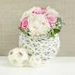 An elegant floral arrangement composed of ivory peonies, fuchsia-pink roses, and soft clouds of baby's breath sits in a modern white woven vase, poised on a clean white tabletop with a neutral beige backdrop that could easily belong to a stylish Highgate apartment. The bouquet is carefully shaped into a compact, rounded form, reminiscent of a bridal bouquet resting for a moment before the walk to a nearby church or registry office. The peonies are the stars, their creamy petals opening in generous layers that create a sense of depth and softness, like finely folded fabric. Surrounding them, the roses glow in shades of vivid pink, their petals tightly clustered at the centres and gently unfurling towards the edges, giving each bloom a sense of movement and life. Delicate sprigs of Gypsophila thread through the composition, their tiny white flowers forming a gossamer-like veil between the larger blossoms, adding a romantic, almost misty quality. Smooth, broad green leaves arc around the base of the bouquet, hugging the flowers and providing contrast with their rich, polished surfaces. The vase is crafted from slender white strands, interlaced in an open, cage-like structure that feels both handcrafted and contemporary, letting the stems show through and casting soft shadows on the table. In front of the vase, two decorative orbs made from pale, woven material echo its texture and add a subtle, sculptural accent. The overall mood is one of serene celebration, ideal as a wedding or anniversary centrepiece for homes near Highgate Woods or as a refined floral gift delivered across N6, capturing the quiet elegance and leafy calm that characterise the local area.