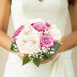 Romantic peony and rose bridal bouquet in ivory, blush pink, and soft lavender, photographed as though poised for a wedding in Highgate's quiet back streets. The bouquet is tightly gathered into a graceful, domed shape that sits snugly in the hands, suggesting a hand-tied design by a skilled local florist rather than a loose, garden-style arrangement. At the heart of the piece are full, creamy peonies, their rounded forms and intricate, overlapping petals giving the impression of tiny clouds or billows of silk, each bloom rich with delicate texture. Encircling them, roses in varied shades of soft pink, warm mauve, and gentle violet add refined structure and depth, their petals spiralling inward toward shaded centres. Threaded finely throughout are stems of gypsophila, their miniature white blooms forming a veil-like halo that softens any sharp edges and adds a classic bridal touch, much like lace. Deep, emerald-green leaves emerge around the lower edge, providing a subtle frame that enhances the pastel palette and introduces a fresh, natural energy reminiscent of tree-lined avenues near Highgate Wood. The lighting appears diffuse and natural, revealing every ruffle and curve without stark shadows, as if the bouquet is resting on a table near a sunlit window in a Highgate home. This arrangement feels suited to an elegant yet understated wedding day, for a bride who appreciates tradition but prefers a clean, modern finish. It evokes calm happiness, tenderness, and a sense of private celebration, like an intimate gathering after vows have been exchanged in a small Highgate chapel.