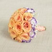 Balanced on a soft, neutral beige backdrop, this bridal bouquet is a radiant symphony of warm peach and gentle orange roses gathered into a lush, domed arrangement. The roses' full blooms invite admiration, promising the quiet luxury often associated with Highgate's historic homes. Among their soft petals, delicate white lisianthus with vivid purple edges add a painterly accent, echoing the creative atmosphere found in Highgate's artful streets and gardens. Sprigs of baby's breath emerge throughout, contributing an airy, ethereal feel that calls to mind the cool morning mist over nearby parks. Bound with a peach satin ribbon and twined with classic pearls, the handle conveys timeless grace and comfort. Ideal for North London bridal parties, celebratory anniversaries, or as a luxurious gift, this bouquet's harmonious palette and elegant finish make it a perfect centerpiece for special occasions across the Highgate area, where tradition and vitality bloom side by side.