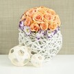 A sophisticated floral display rests on a flawless white surface, illuminated by gentle, even light that makes the colours appear soft and inviting, much like a tranquil interior near Highgate Woods. At the heart of the image is a dome of roses in shades of peach, apricot, and light salmon, carefully arranged into a smooth, spherical shape. Each rose is fully open, revealing concentric layers of velvety petals that overlap and fold into each other, creating a sense of fullness and generosity. Encircling the base of this rose globe is a ring of lisianthus flowers, their white petals delicately frilled and outlined with a bold band of violet or deep purple, adding contrast and visual rhythm. Between these blooms, tiny sprays of white gypsophila appear, their fine clusters of blossoms reminiscent of tiny stars scattered through a mist, lending an airy quality that lightens the look. The arrangement is set in a distinctive rounded vessel woven from white wicker or rattan, its open lattice design forming a bulbous, sculptural basket. The weave allows light to pass through and cast intricate shadows on the bright surface below, subtly highlighting the craftsmanship. In front of the main arrangement, two decorative spheres made from pale, natural wooden strips rest casually; one is noticeably larger than the other, and both echo the open, woven motif of the container. Behind, a delicately textured beige fabric backdrop provides a warm, neutral setting that lets the floral colours and textures stand out. The mood of the piece is romantic and serene, perfectly suited to wedding receptions, anniversaries, or elegant Highgate homes seeking a touch of natural, contemporary charm, with Flowers Highgate capturing that balance of softness and structure.