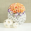 This photograph presents a thoughtfully curated centrepiece where artistry and local sensibility meet: approximately two to three dozen apricot and soft peach roses form a domed mass, their petals layered like silk and cinnamon, some open to reveal fragrant hearts, others curled into tender buds. White Lisianthus ring the roses, their delicate throats painted with vivid indigo-purple along the petal edges, creating a refined contrast that catches the eye and lifts the palette. Scattered throughout are small sprays of fine white Gypsophila (baby's breath), providing a light, cloud-like veil that adds breathability and a timeless bridal feel. The flowers rest in a contemporary white woven orb - an open-lattice vase of reed or rattan that feels both handmade and modern - its bright surface reflecting the soft daylight. Beside the arrangement on the smooth, clean white surface are two woven wooden spheres, one larger and one smaller, their pale textures mirroring the basket and adding an artisanal decorative touch. A softly textured beige-linen wall forms a muted backdrop, gently out of focus so the bouquet's tones and textures remain central. The composition reads as romantic and sophisticated, perfect for wedding decor, anniversary gifts, or a refined home centrepiece. As a local florist in Highgate, Flowers Highgate might prepare this arrangement for a village hall reception near Pond Square or for a relaxed table at a Hampstead Heath picnic, with options for local delivery and careful handling to preserve the fragrance and form.