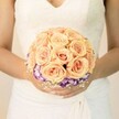 A softly lit, chest-level photograph centres on a bride standing very still, her hands gently cupping a round bridal bouquet as if it were the quiet heart of the day. She wears a classic white or ivory wedding gown with a lace bodice that traces delicate patterns across the fabric, the neckline forming a subtle V that feels both modest and romantic. The skirt falls in smooth, graceful folds, catching the light in gentle ripples, like the way sunlight brushes across the lawns on a calm morning in Waterlow Park. Nestled against the gown, the bouquet glows with warm, intimate colour. At its core are tightly clustered roses in shades of peach, apricot, and blush pink, each bloom full and velvety, the petals spiralling inward to deep, romantic centres. Their tones suggest early evening light over Highgate Village, soft but confident, filled with anticipation. Beneath this cloud of roses runs a band of deep purple blooms edged in creamy white, likely lisianthus, their ruffled petals adding a ring of contrast that frames the roses with a touch of drama. Tiny sprays of white filler flowers, reminiscent of baby's breath, weave around the base like a fine lace cuff, echoing the gown's detailing and adding airiness and movement. Her nails are subtly manicured in a pale shade, with one thumb catching a hint of shimmer, like a discreet glint of city light along Highgate Hill after dusk. The overall mood is calm, romantic, and personal-a timeless bridal bouquet that might be carried from a ceremony in a Highgate church to a reception nearby, a floral companion to vows spoken softly and remembered for years.