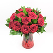 A studio-style photograph shows a classic bouquet of deep red roses arranged in a rounded dome and placed in the centre of a stark white background, so every detail of the flowers stands out. Around a dozen velvety crimson roses, each bloom fully open with spiral centres and softly curled outer petals, are grouped closely together to create a sense of abundance and romance. Between the roses, polished dark green leaves, similar to Ruscus foliage, peek through and edge the base of the bouquet, their glossy texture catching the light and giving the arrangement structure and freshness. The bouquet rests in a translucent ruby-red glass vase with a gently flared rim and base; faint vertical swirls or ribbing in the glass create a subtle pattern that glimmers where the light touches it. Through the tinted glass, the straight rose stems can be seen crossing and layering, adding depth and a natural, organic line beneath the precise top of blooms. The lighting is bright and even, much like a clear morning over Highgate Wood, revealing every shadow in the petals and every highlight on the vase without distraction. The mood is unapologetically romantic, ideal for Valentine's Day, an anniversary dinner in a Highgate village townhouse, or a quiet gesture of devotion delivered to a home off Archway Road. This single vase of roses feels confident and passionate, the kind of arrangement a skilled florist would prepare when words feel too small, allowing the rich red flowers and their timeless symbolism to speak clearly in any Highgate setting.