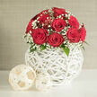This image showcases an elegant, contemporary floral arrangement that would feel right at home on a wedding table in Highgate or in a thoughtfully styled townhouse off Swains Lane. At the centre is a domed bouquet of deep red roses, their petals thick, velvety, and flawlessly formed. Each rose appears at peak bloom, with a compact spiral in the middle and broader outer petals that curl gently downwards, creating a lush, full appearance. Between the roses, sprigs of white gypsophila weave in and out, their tiny clustered flowers resembling soft foam or fine lace. The baby's breath adds a delicate, airy texture and a luminous contrast against the intense crimson petals, giving the whole arrangement a sense of lightness and romance. A few dark green leaves peek discreetly from the base of the bouquet, offering hints of natural foliage without disturbing the carefully rounded silhouette. Supporting this floral dome is a round, sculptural vase crafted from slim, white rods arranged in a woven lattice. The structure, which resembles an artistic wicker or rattan sphere, has open gaps that allow light to filter through, casting faint, geometrical shadows on the bright white tabletop below. Beside the vase, two decorative balls made from similar woven material sit in relaxed harmony, their cream or off-white tones subtly echoing the vase's design while keeping the focus firmly on the flowers. The background is a softly textured wash of neutral beige or light grey, undramatic and calm, allowing the bouquet's colours and textures to dominate. Soft, balanced lighting bathes the arrangement, highlighting the velvety sheen of the roses and the fine detail of the gypsophila, creating an impression of refined romance and careful craftsmanship, ideal for wedding collections, anniversary surprises, or special celebrations across N6.