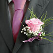 Pinned neatly to a charcoal jacket, this refined boutonniere features a single blush pink rose that seems to glow against the depth of the suit fabric, quietly capturing the mood of a wedding morning in Highgate. The rose is in ideal condition, gently unfurling with layers of velvety petals that swirl inward to a softly blushed centre, suggesting tenderness and new beginnings. Around it float airy clusters of white gypsophila, each tiny star-shaped flower contributing to a gauzy, cloudlike effect that lightens the composition and creates a lovely contrast with the dark lapel. Slender, arching strands of fresh greenery frame the rose, adding a sense of natural movement reminiscent of the foliage around Highgate Wood and the green edges of nearby streets. The stems are carefully bound with a pale lilac or muted mauve satin ribbon, tied into a small, precise bow that sits just beneath the bloom, offering a hint of sheen that echoes the subtle shine of a silk tie. The lighting catches the rose at just the right angle, emphasizing its softness while defining the crisp lines of the suit, making each detail stand out clearly for photographs. This boutonniere feels ideal for a groom, best man, or prom guest heading out from a local Highgate florist, carefully chosen to complement a classic suit without overshadowing it. There is a sense of calm anticipation in the design, as though the wearer is moments away from stepping into a ceremony, greeting guests, and creating memories that will remain as vivid as the rose's colour long after the petals have fallen.