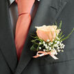 A single peach rose sits serenely on a charcoal lapel in this carefully balanced boutonniere, its petals layered like soft porcelain and warming from pale blush at the outer edges to a deeper coral-pink in the center. Encircling the rose, tiny white gypsophila sprigs create a frothy, cloud-like texture; their clustered, starry blooms add a whisper of innocence and light contrast that makes the peach hue read brighter against the suit. Sprays of fine green foliage provide structure and a fresh counterpoint-slender, slightly serrated leaves that lend a hint of garden-fresh aroma and suggest early-morning dew. A satin ribbon, closely matched to the tie's coral tone, is tied into a compact bow at the buttonhole, its subtle sheen mirroring the tie's soft luster and pulling the palette together with quiet precision. The charcoal fabric of the jacket acts as a neutral, elegant canvas, deepening shadows around the floral elements and highlighting the tactile differences between the rose's velvety petals, the gossamer of the gypsophila, and the crispness of the foliage. The overall effect is composed and refined-ideal for ceremonial occasions where subtlety matters-conveying both celebration and thoughtful care. Imagine this boutonniere travelling from a Highgate workshop toward Pond Square or North Hill, where the neighbourhood's calm streets and leafy corners make such restrained, tasteful details feel right at home as part of a wedding look or a formal anniversary outfit.