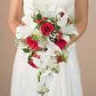 In this close-up photograph, the camera lingers lovingly on a dramatic wedding bouquet, held securely at the bride's waist so that her white, delicately textured gown frames the floral cascade, evoking a scene from a Highgate village ceremony. The bouquet itself is composed in a waterfall style, with a generous cluster of deep crimson roses at the top. These roses are plush and velvety, each bloom rich with layered petals that spiral towards the centre, giving the arrangement a sense of warmth and passionate intensity. Nestled between them, pure white lilies open in generous, star-shaped blossoms, their firm, porcelain-like petals curling slightly at the tips and revealing subtle green throats and stamens. From this central cluster, stems of white Phalaenopsis orchids sweep downward, creating a graceful cascade. Their petals are smooth and luminous, with a slight sheen that catches the light, and the mix of fully opened flowers and tight buds adds dimension and gentle movement. Fine sprays of baby's breath lace through the bouquet, their tiny white blooms forming an airy veil that softens the strong red-and-white contrast. Interspersed are bright red hypericum berries, rounded and glossy, introducing a hint of playful texture and a festive touch. Around and behind the flowers, a variety of lush greenery-wispy, fern-like fronds, slender leaves, and deeper green accents-provides depth, structure, and a sense of fresh vitality. The background recedes into a smooth, neutral blur, allowing the intricate textures and colours of the blooms to stand out. Designed with the discerning Highgate bride in mind, this bouquet feels ideal for weddings near Highgate Wood, local churches, or intimate townhouse receptions.
