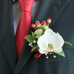 Perched on the left lapel of a tailored charcoal jacket, this boutonnière reads as a small, composed story of texture, colour, and careful hands. The central element is a white Phalaenopsis orchid whose broad, velvety petals open with an almost luminous calm, delicate at the edges and cool to imagine to the touch. Nestled within the bloom, a yellow-gold patterned labellum offers a faint, intimate accent that lifts the composition. Glossy, deep-green foliage surrounds the orchid like a soft frame, grounding the arrangement in natural weight and fresh scent. Scattered sprigs of baby's breath lend an airy, nostalgic aura - tiny pearls of white that soften the silhouette and whisper of hopeful beginnings. Clusters of pink-to-coral hypericum berries introduce a rounded rhythm and a confident pop of red, harmonising with the solid red satin tie and the subtle sheen of the matching ribbon looped at the boutonnière's base. A light-coloured, subtly patterned shirt shows beneath the lapel, its grid-like texture providing a quiet contrast to the suit's dark tone and the tie's vivid colour. Soft, even lighting captures the orchid's gentle translucence and the berries' glossy highlight, making the ensemble feel both celebratory and intimate. Ideal for weddings or milestone evenings in and around Highgate Village or a riverside reception after a stroll through Hampstead Heath, this boutonnière reflects considered floral craft and the calm confidence of wearing something made with care.