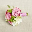 A small, romantic bouquet lies horizontally on a softly textured, light tan fabric, the weave of the cloth gently catching the light like a linen runner on a Highgate dining table. At the heart of the arrangement, two fully opened pink roses and a slightly smaller third bloom draw the eye, their petals shaded from blush to deeper magenta at the frilled edges, creating a subtle ombré effect. Each rose is lush and layered, the centres still slightly furled, suggesting that the flowers were cut fresh, perhaps that same morning. Around them sit rounded tufts of lime-green button chrysanthemums, their tiny, tightly packed petals forming playful, pom-pom spheres that contrast beautifully with the softness of the roses. Creamy white filler flowers, likely lisianthus or freesia, tuck in between the main blooms, their delicate, papery petals adding volume and a sense of airiness. Smaller starry white blossoms with yellow centres glimmer here and there, like scattered confetti after a ceremony on Highgate Hill. The stems are gathered and neatly bound into a compact posy; a bright fuchsia satin ribbon wraps the handle with precision, its surface smooth and light-catching. Over the ribbon, a fine strand of white faux pearls winds diagonally, as though a bracelet has been lovingly fastened around the bouquet, leaving the trimmed green stems to peek out below. The lighting is gentle and even, with no harsh shadows, lending the flowers a calm, flattering glow, as if they are waiting on a table at a local Highgate pub prepared for a wedding reception. The overall impression is of a chic, intimate bridal or bridesmaid bouquet, perfect for a registry office ceremony or small celebration, its classic pink, green, and white palette conveying fresh romance and understated elegance.