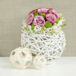 On a sleek white tabletop that subtly reflects its shapes and colours, a beautifully crafted floral centerpiece creates a sense of poised celebration, reminiscent of a quiet wedding breakfast in Highgate. The arrangement is anchored by a spherical vase made from white-painted natural twigs, intricately woven into a light, open lattice. Each curved branch forms part of a delicate network, giving the container an airy, nest-like quality that feels both organic and distinctly modern, perfect for stylish receptions near Highgate Woods or contemporary ceremonies in local venues. Emerging from this textured globe is a rounded dome of flowers, meticulously arranged for balance and harmony. At its heart, soft pastel pink roses open in luxuriant, full blooms, their velvety petals spiralling outward and, in some, deepening to a richer pink at the edges. Between these roses, bright lime-green button chrysanthemums nestle like small jewels, their compact, spherical forms adding playful texture and a refreshing burst of colour. Around the base, a fringe of small white flowers-likely lisianthus or stock with pale yellow centres-spills gently over the rim, softening the line between the floral canopy and the structural weave of the vase. To the left, two pale wooden orbs, one larger and one smaller, rest on the tabletop, composed of thin strips that twist and interlace, echoing the same openwork pattern and enhancing the rustic-chic theme. Behind the scene, a warm, neutral background reminiscent of woven fabric or softly brushed plaster is intentionally blurred, allowing the details of each flower and twig to stand out clearly. The overall effect is of serene elegance and gentle joy, ideal for wedding tables, anniversary dinners, or engagement celebrations across Highgate and the surrounding North London neighbourhoods.