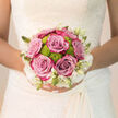 This image captures a bride holding a carefully crafted bouquet, the blossoms gathered close to her dress as though she is drawing in a little extra courage and happiness before the ceremony. At the centre are generous roses shaded between lilac and dusty pink, their petals layered in soft, rounded spirals that make you think of early evening skies above Highgate's rooftops. Between these roses sit bright lime-green button chrysanthemums, neatly rounded and densely petalled, creating a playful rhythm of colour and a contemporary twist within the otherwise timeless arrangement. Encircling the bouquet, creamy white blooms and fine buds form a soft halo, like delicate lace tracing the edges of a bridal veil, or the pale blossoms that appear in springtime around Pond Square. Gentle, natural light falls across the flowers, highlighting each curve and texture without harshness, suggesting a calm moment in a Highgate home or boutique venue just before guests arrive. In your mind's nose, you might detect a subtle fragrance: the familiar, comforting scent of roses carefully balanced with the cool, green aroma of freshly cut stems. The stems are bound in a secure, tidy handle, allowing the bouquet to rest snugly in the bride's hands as she moves from ceremony to photographs, perhaps pausing near the gates of Highgate Cemetery or a quiet corner of Waterlow Park. The overall mood is intimate and quietly hopeful; this is a bouquet that feels thoughtfully designed, ready to stay fresh and radiant throughout a day filled with promises and celebration.
