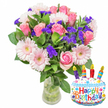 Bathed in soft morning light against a clean white backdrop, this celebratory bouquet radiates a gentle, joyful energy perfect for a birthday. At its heart are dozens of soft pink roses, petals unfurling from palest blush to a warmer, deeper pink, their velvety texture catching the light and suggesting a classic romantic warmth. Interspersed like cheerful punctuation are large pale-pink Gerbera daisies, their layered petals opening wide and centring on a buttercream-yellow eye, adding a playful, bright note. Scattered through the composition are clusters of vivid purple filler flowers-statice or limonium-with a papery, multi-faceted texture that gives the arrangement depth and a touch of wild meadow character. Lush, glossy deep-green leaves cradle the blooms and provide a cool backdrop that makes the pinks and purples sing, while a clear glass vase with a slightly wider base reveals the tangled green stems and fresh water below, reinforcing the impression of recently gathered, hand-tended blooms. To the lower right, a whimsical cake-shaped Mylar balloon punctuates the scene: a white frosted cake motif edged with wavy blue piping, sprinkled with red, yellow, blue and green dots, and topped with four striped candles-purple, red, orange and blue-each capped with a tiny yellow flame. Across the front, the words Happy Birthday appear in playful, multi-coloured letters, and a small KALEIDOSCOPE logo sits unobtrusively at the balloon's base. This image reads as a thoughtful birthday gift - a florist's composition that balances softness, celebration, and freshness - suitable for local delivery around Highgate, Waterlow Park and the village green.